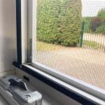 Close-up of Fliegengitter Fenster - mit Rahmen with latch, showing greenery and a gravel path outside.